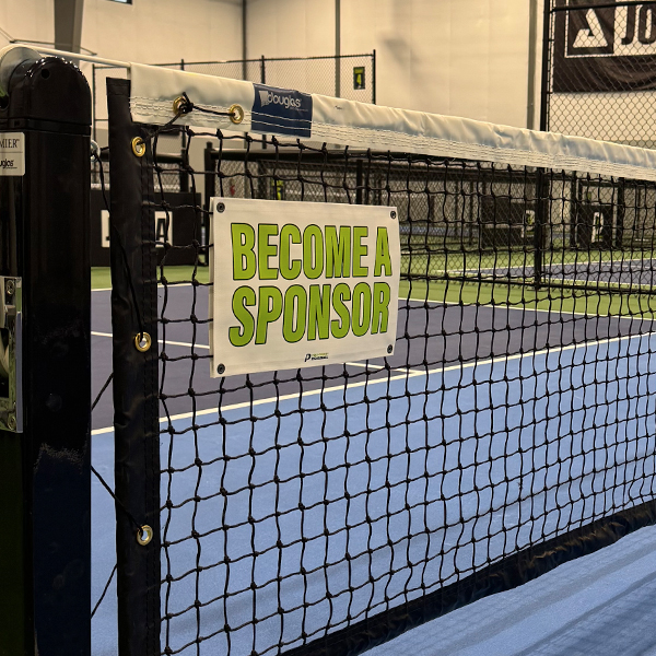 White 'Become a Sponsor' net banner with bold green lettering snapped onto an indoor pickleball court net